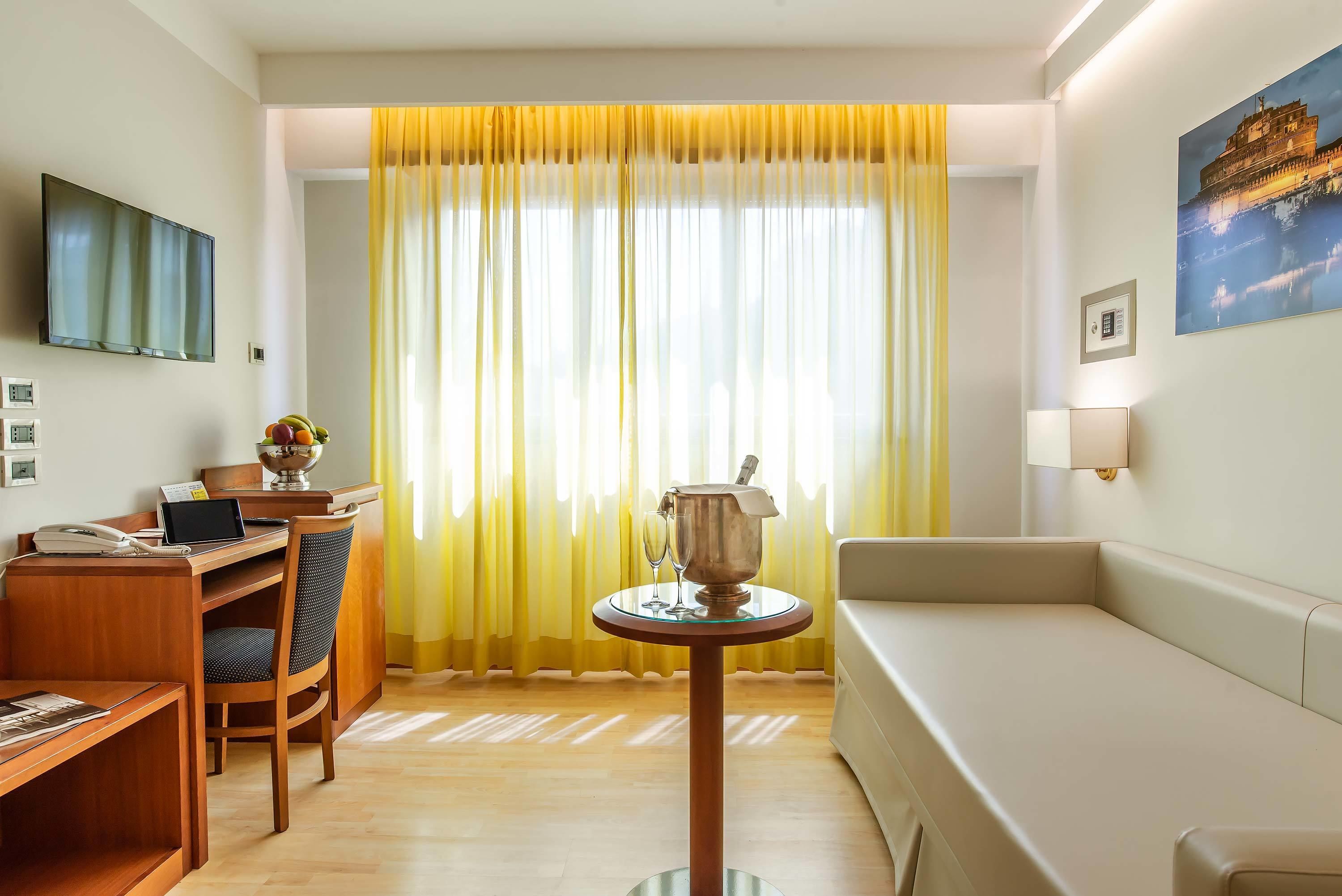 A hotel room with a wooden desk, a laptop, a telephone, a chair, a small round table with a decorative item, a sofa, a television mounted on the wall, and a large window with yellow curtains.