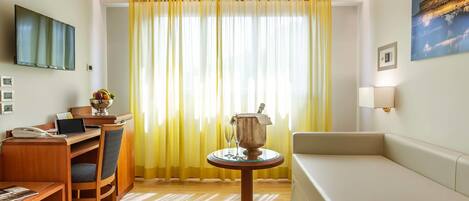A hotel room with a wooden desk, a laptop, a telephone, a chair, a small round table with a decorative item, a sofa, a television mounted on the wall, and a large window with yellow curtains.