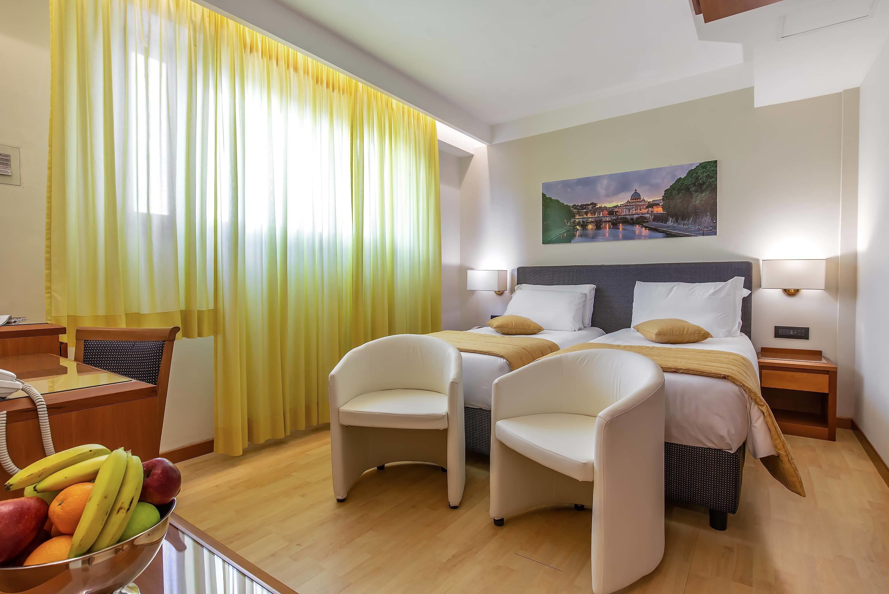 A hotel room with a bed, two chairs, a desk with a phone, and a fruit bowl.