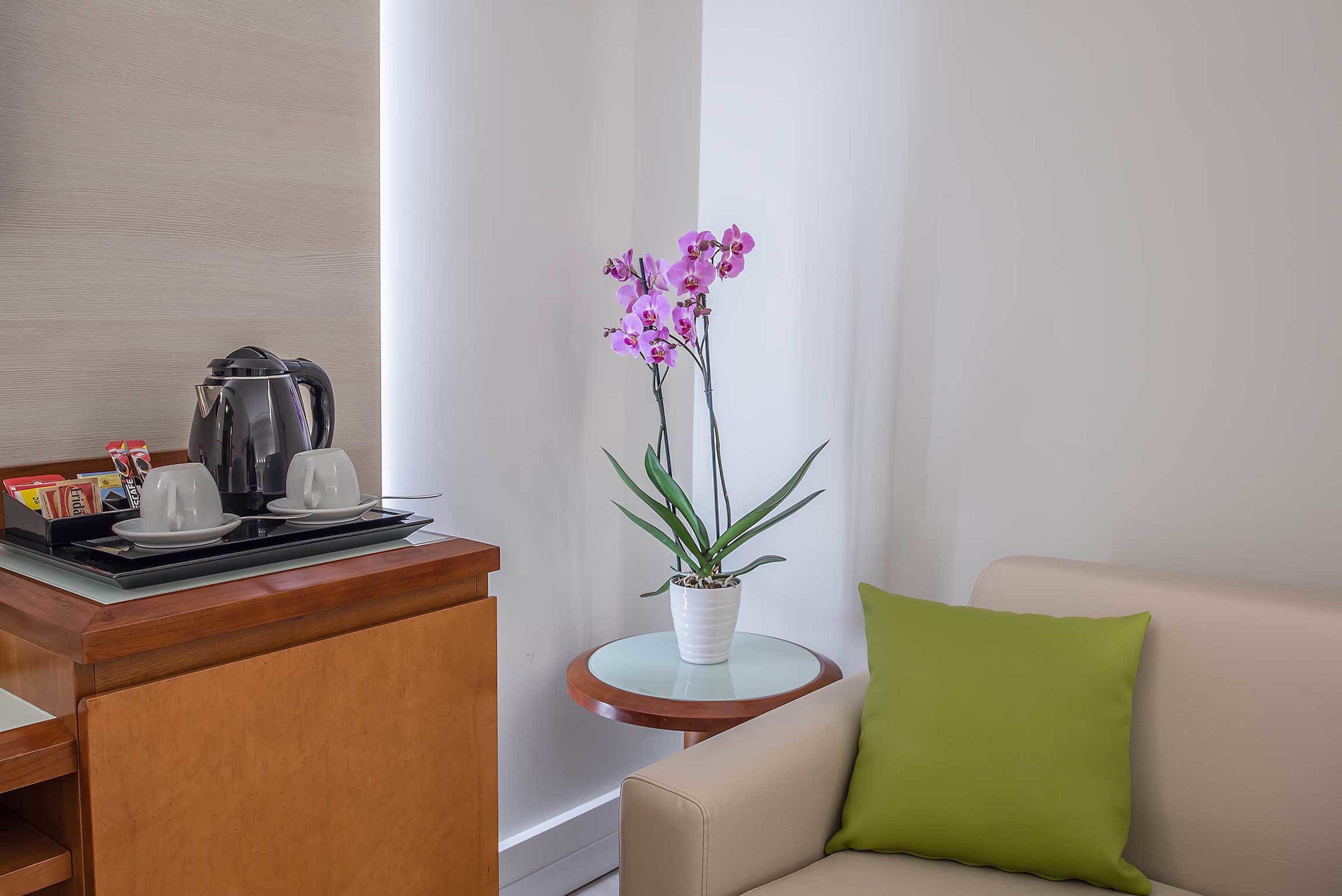 A hotel room with a wooden desk, a coffee maker, a potted orchid, a round table, and a beige sofa with a green pillow.
