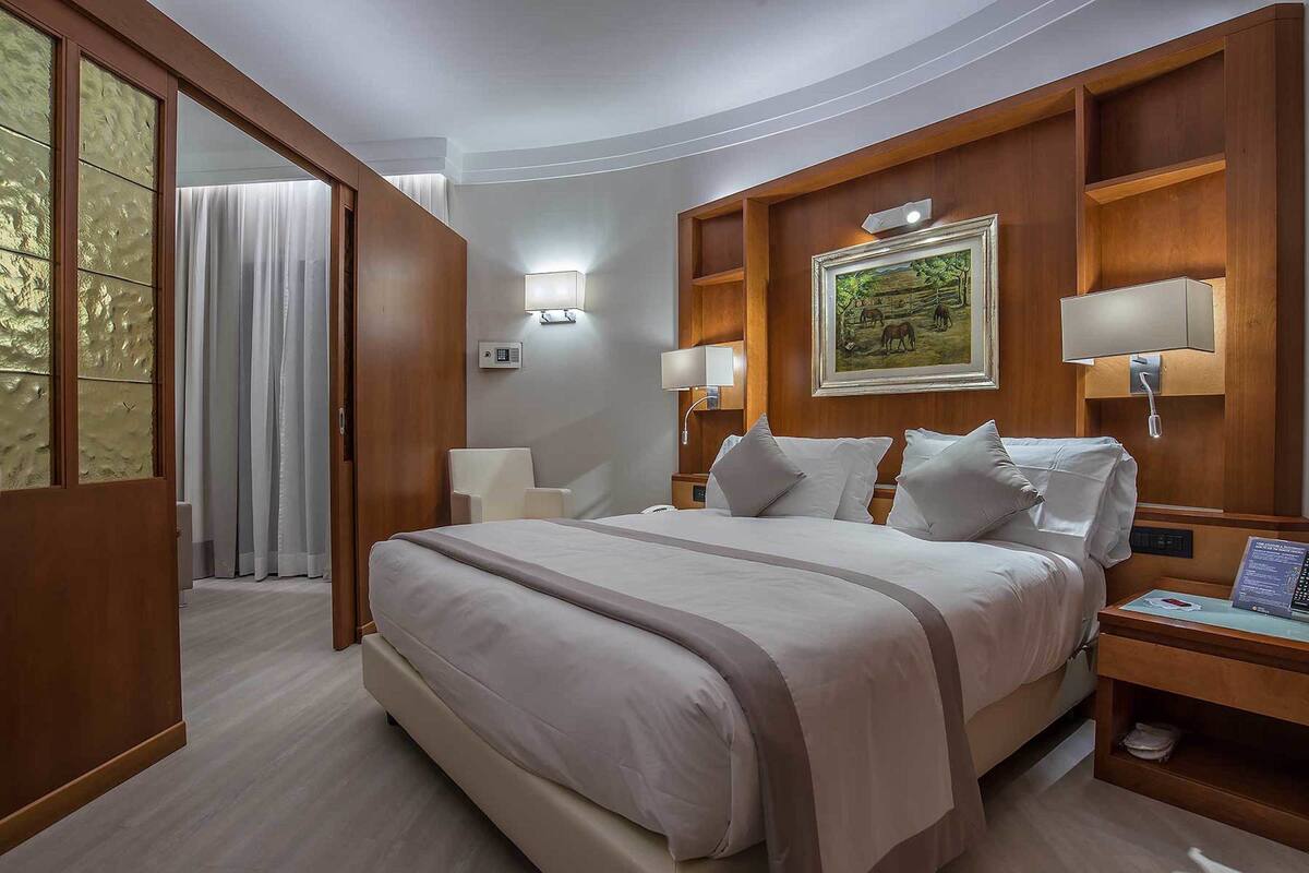 A hotel room with a large bed, wooden headboard, two bedside tables with lamps, a painting on the wall, and a door leading to another room.