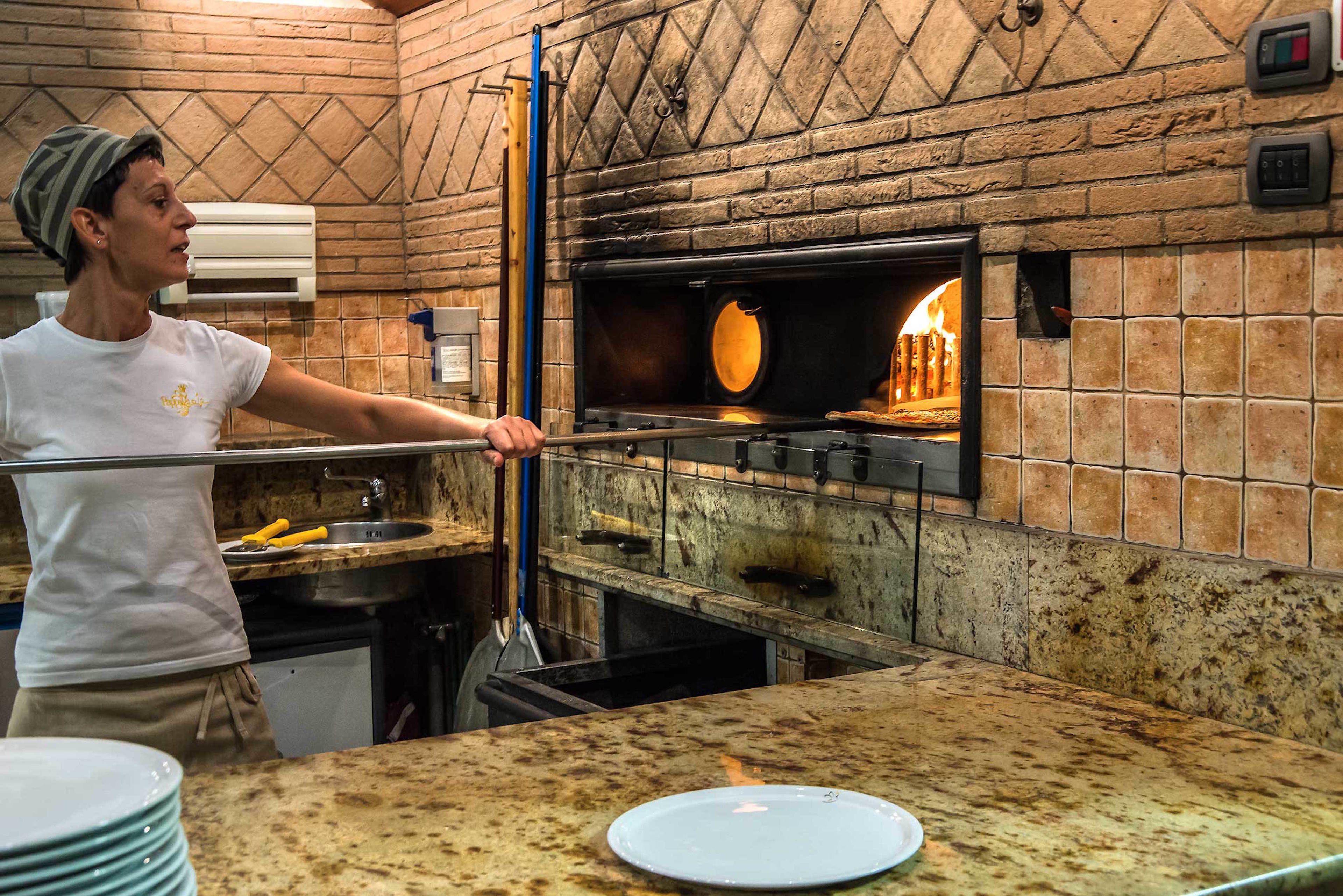 A kitchen with a brick wall, a pizza oven, and a person using a pizza peel.