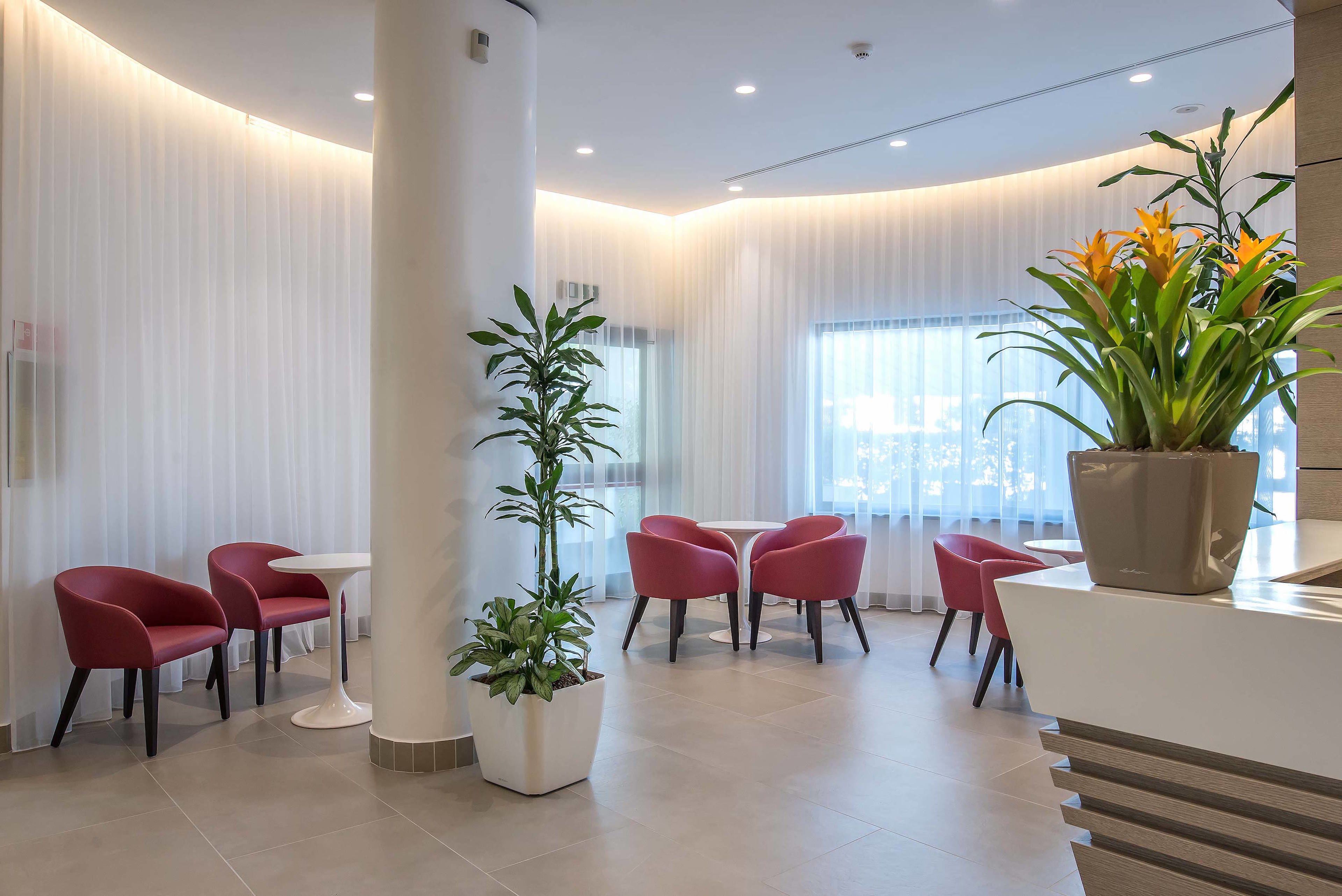 A modern hotel lobby with red chairs, a white reception desk, and large windows.