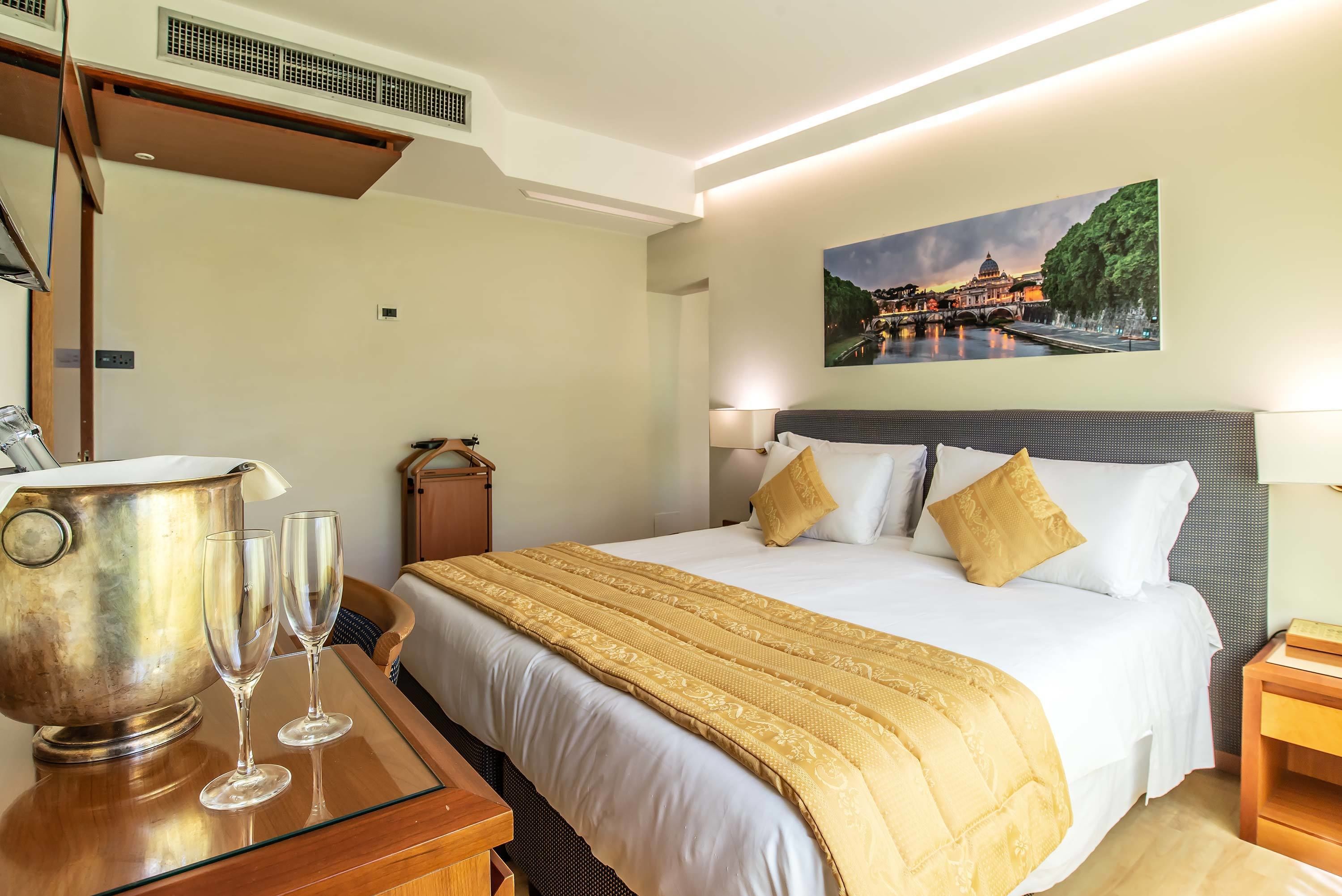 A hotel room with a bed, bedside tables, a nightstand, a wall-mounted painting, and a large metallic object on the floor.