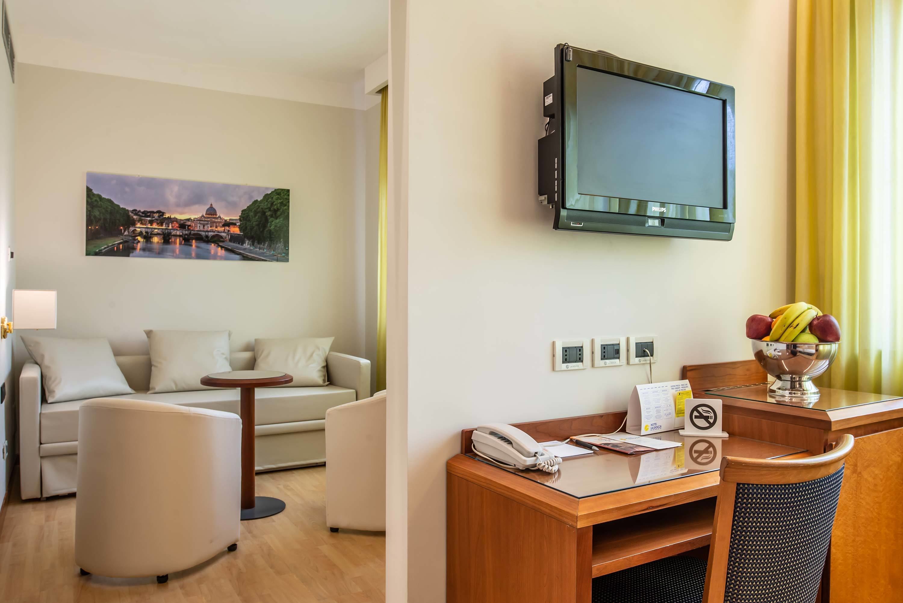 A hotel room with a flat-screen TV mounted on the wall, a sofa, a small round table, a desk with a telephone and a fruit bowl, and a painting of a bridge.