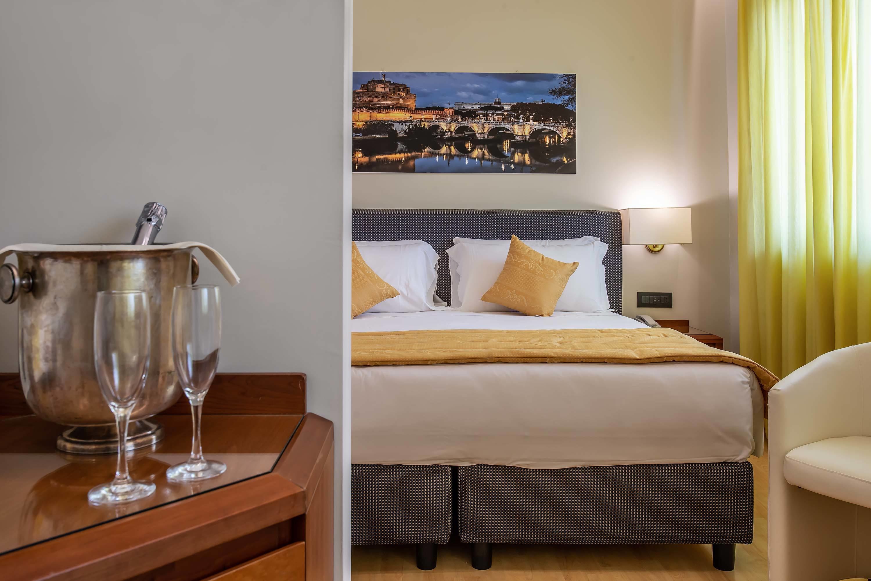 A hotel room with a bed, a nightstand, a lamp, a large painting of a cityscape, and a view of a yellow curtain.