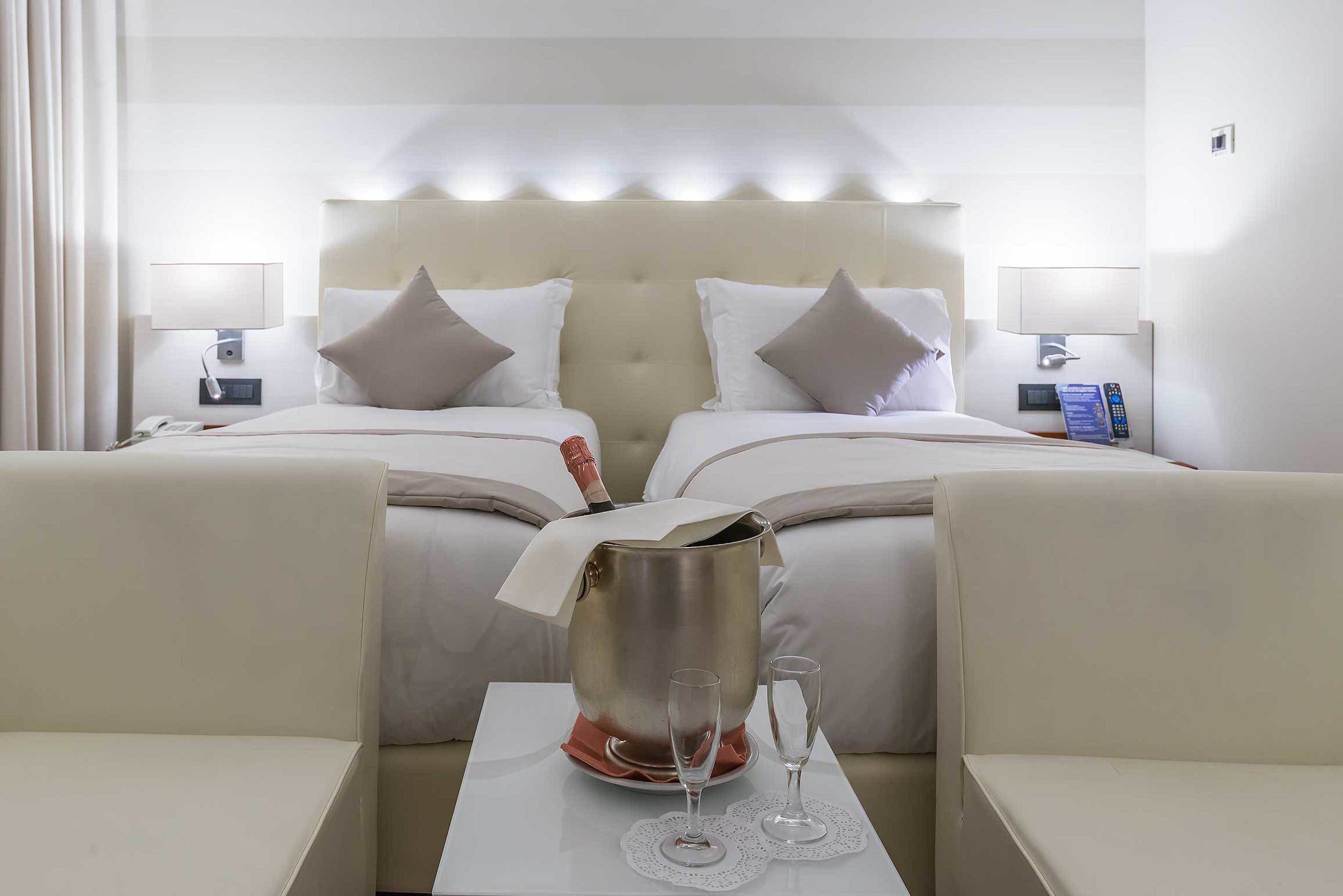 A hotel room with two beds, a minibar, a table with two glasses, and a remote control on the nightstand.