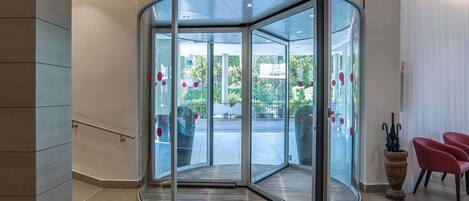A modern hotel lobby with a glass revolving door, a staircase, and a seating area with red chairs.