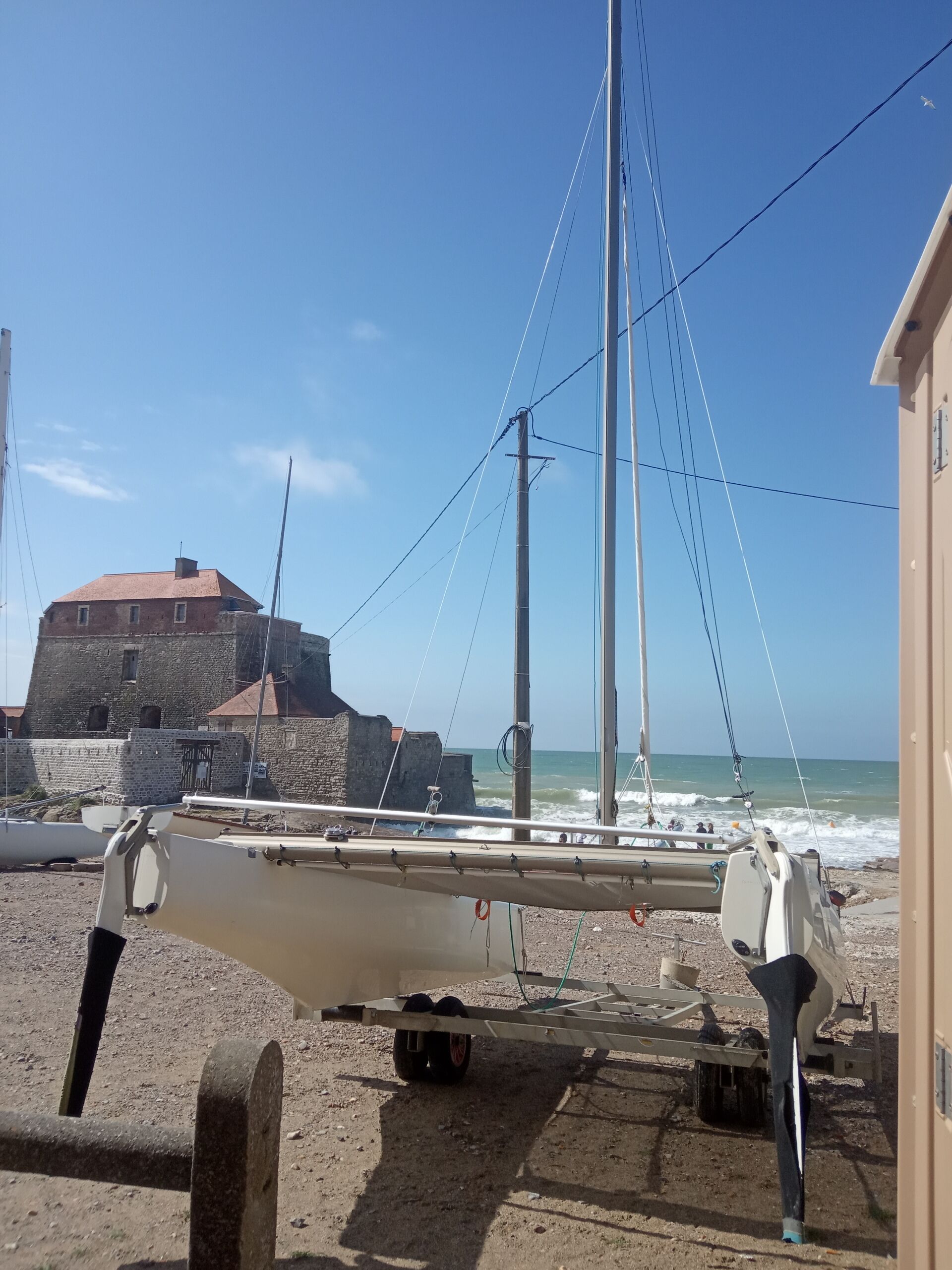 Een zeilboot afgemeerd aan een zandstrand, met een stenen gebouw en een helderblauwe lucht op de achtergrond.