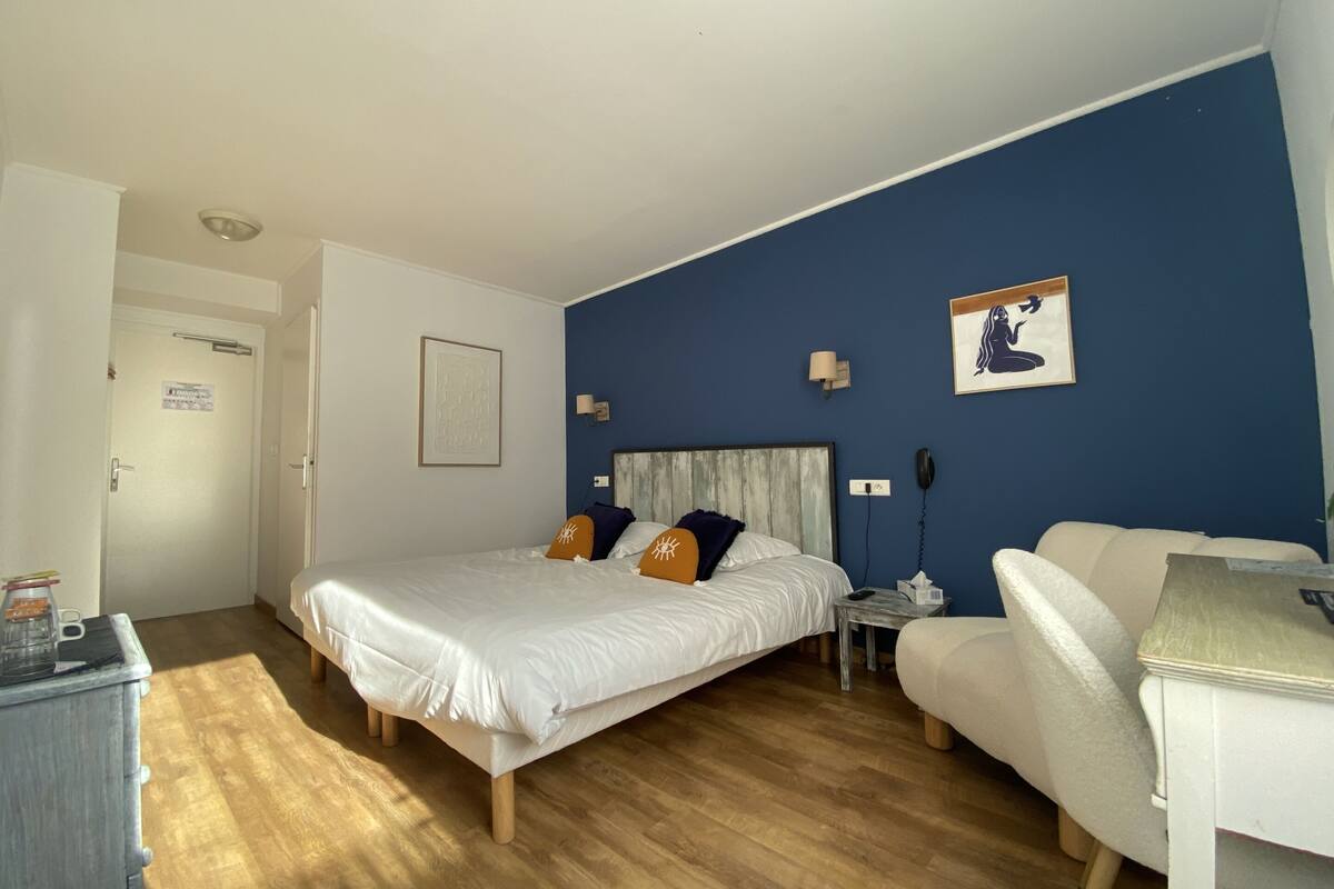 A hotel room with a bed, a desk, a chair, a nightstand, and a wall with a framed picture.