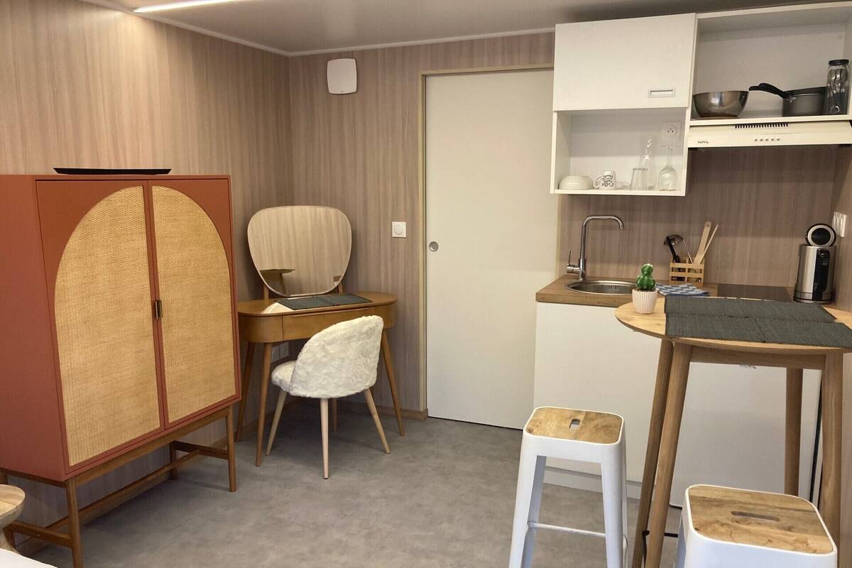 A compact kitchen with a wooden cabinet, a white chair, a small stool, and a dining table.