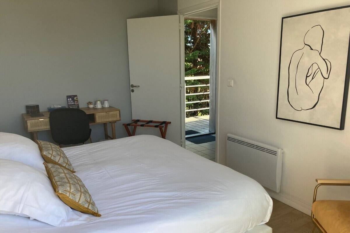 A neatly made bed with white linens and patterned pillows, a desk with a chair, a radiator, and a framed artwork on the wall.