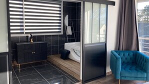 A modern bathroom with a black tiled wall, a white bathtub, a black cabinet with gold handles, a blue chair, and a large window with blinds. - Hôtel des Argousiers (Ambleteuse)