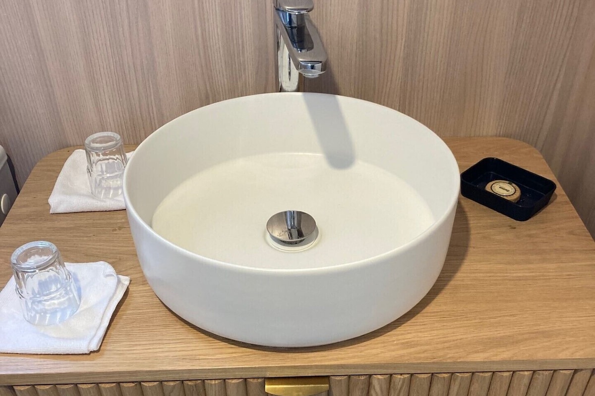 Un baño con un lavamanos blanco y redondo, un grifo, un vaso pequeño y un jabonera.