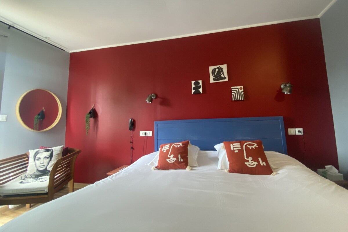 A hotel room with a red accent wall, a blue headboard, and a bed with white bedding and red pillows.