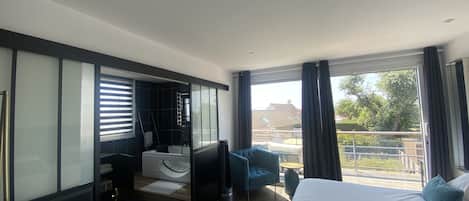 Une chambre d’hôtel moderne avec un grand lit, un coin salon comprenant un fauteuil bleu et une petite table, et une vue sur l’extérieur.