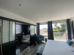 A modern hotel room with a large bed, a seating area with a blue armchair, a small table, and a view of the outside. - Hôtel des Argousiers (Ambleteuse)