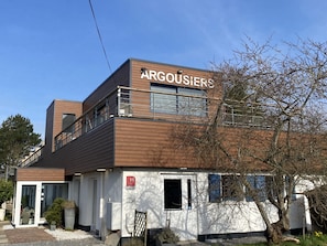 A modern building with a sign reading "ARGOUISIERS." It features a wooden facade, large windows, and a small entrance with a door.