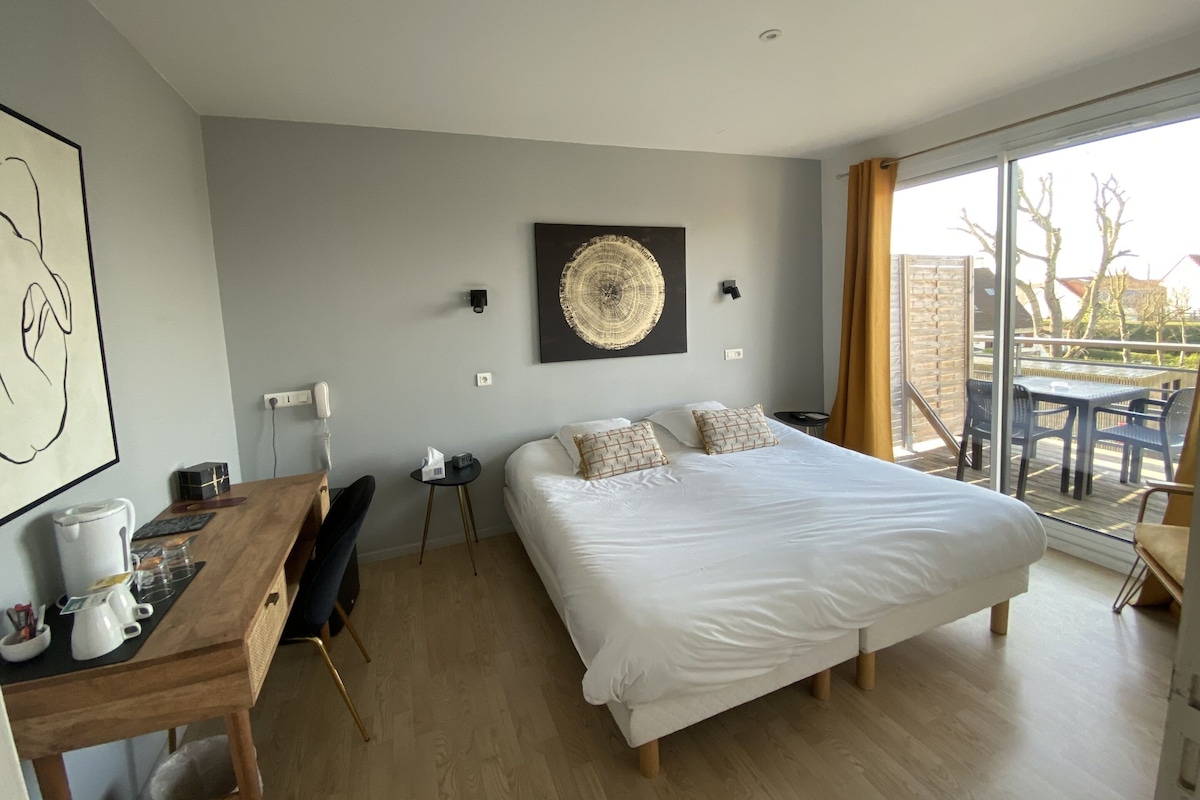A modern bedroom with a large bed, a desk with a kettle, a chair, and a view of the outdoors through a glass door.