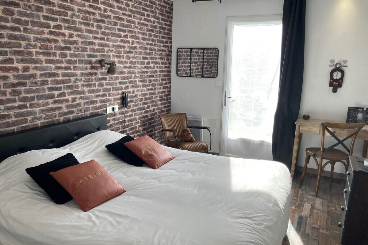 A bedroom with a brick wall, a bed with white bedding and two pillows, a chair, a desk, and a clock on the wall.