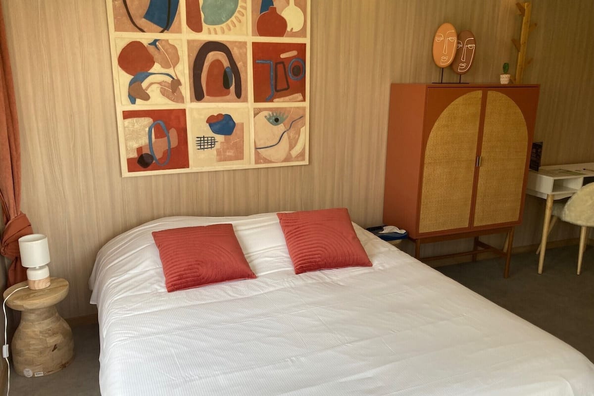 A hotel room with a large bed, two red pillows, a wooden cabinet, a desk, and abstract artwork on the wall.