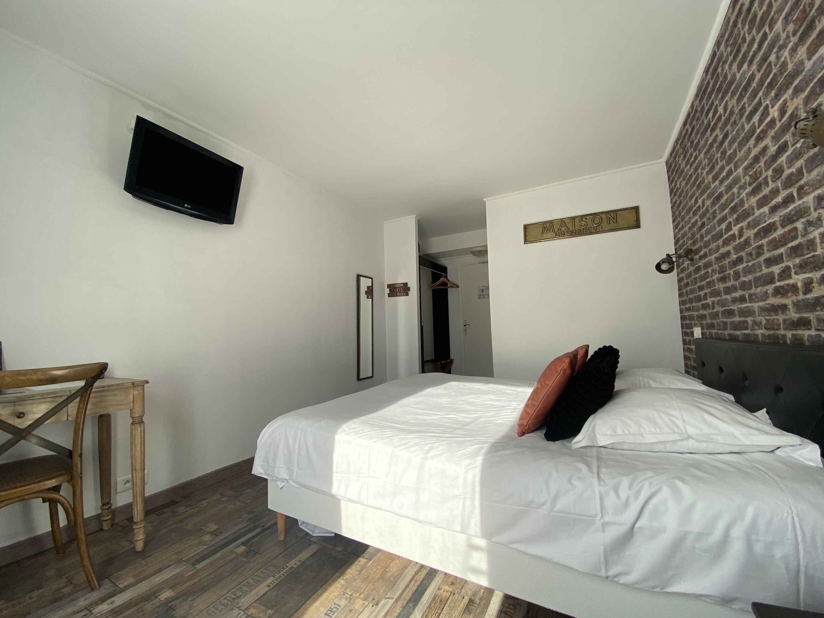 Une chambre d’hôtel avec un lit, un téléviseur fixé au mur, une chaise en bois, un miroir et un mur en briques.