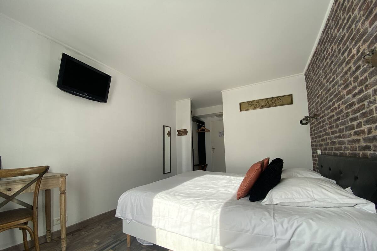 A hotel room with a bed, a TV mounted on the wall, a wooden chair, a mirror, and a brick wall.