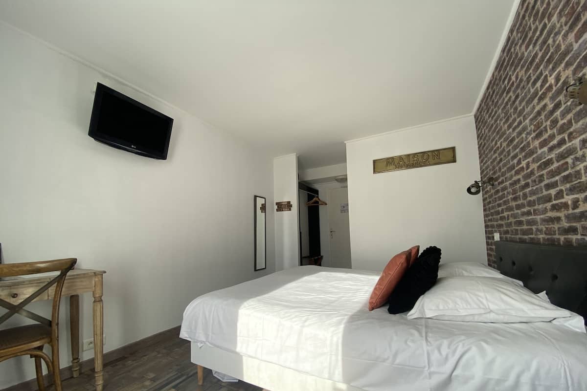 Una habitación de hotel con una cama, un televisor instalado en la pared, una silla de madera, un espejo y una pared de ladrillo.