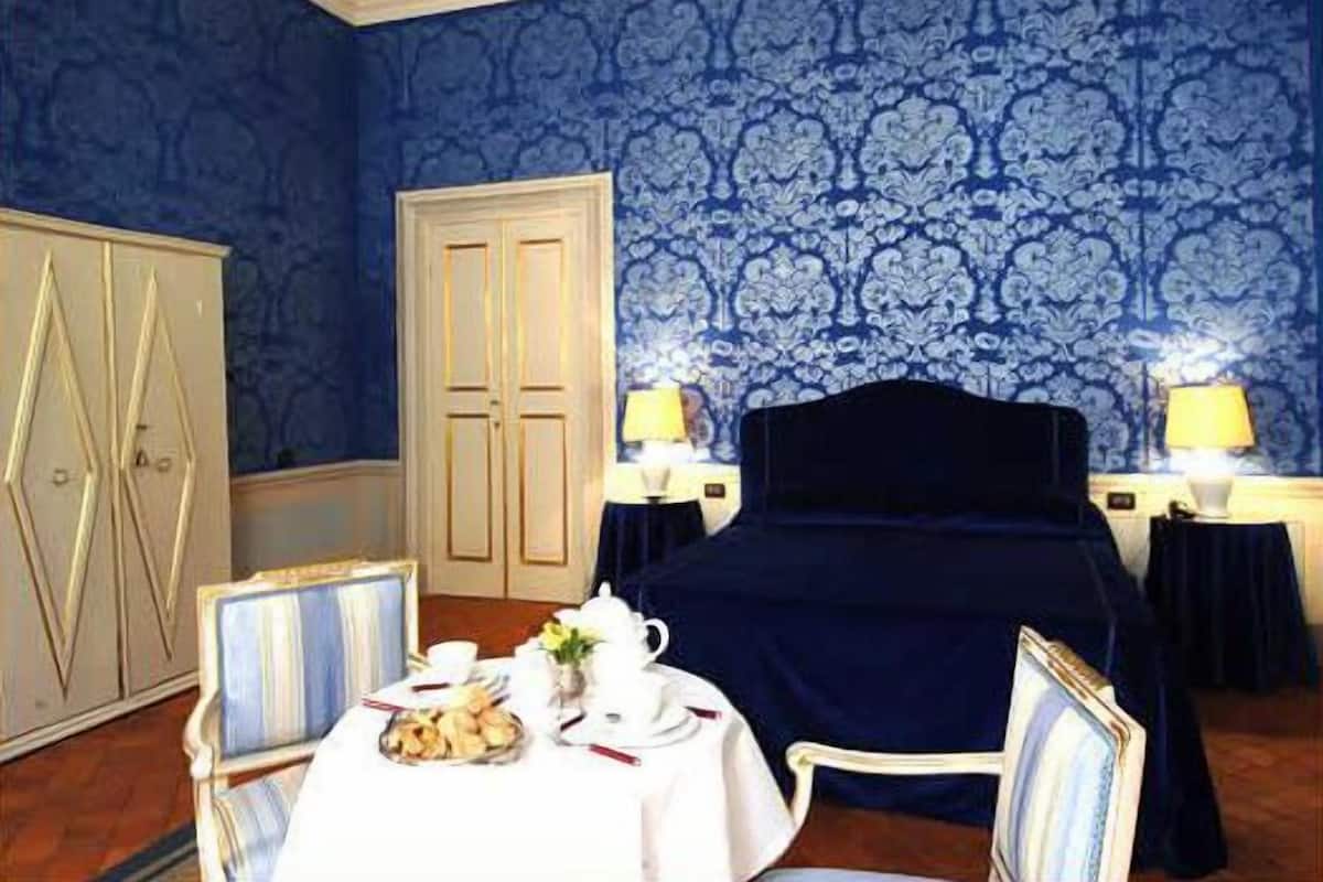 Habitación de hotel con papel tapiz azul estampado, una cama con cubrecama oscuro, una mesa con tetera y pastelería, y dos lámparas de noche.