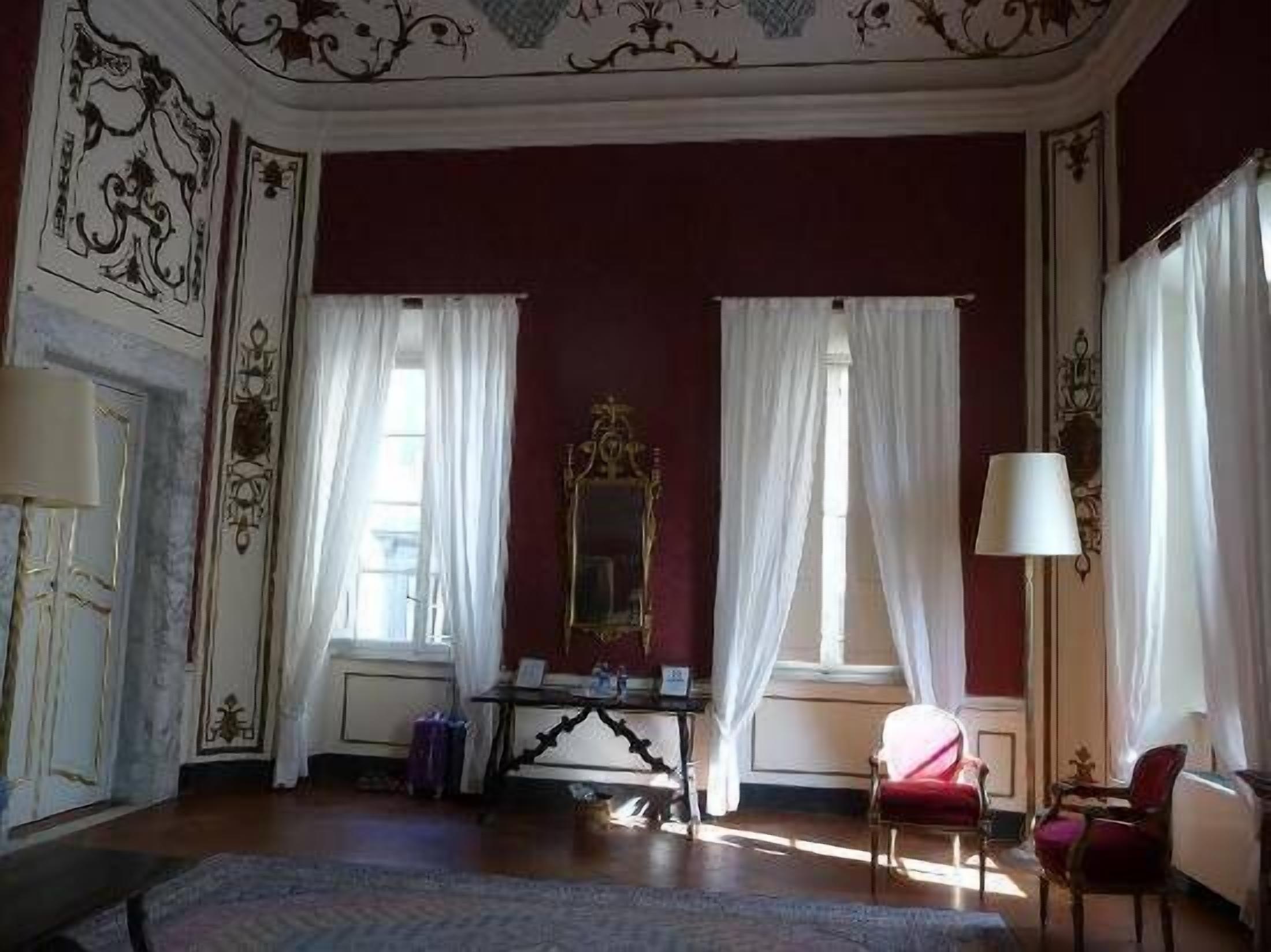 Una habitación con un espejo grande, dos ventanas con cortinas blancas, una silla roja y una mesa con una lámpara.