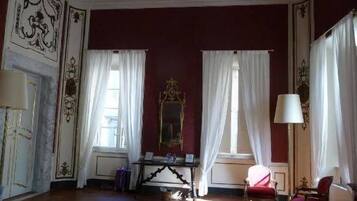 Una habitación con un espejo grande, dos ventanas con cortinas blancas, una silla roja y una mesa con una lámpara.