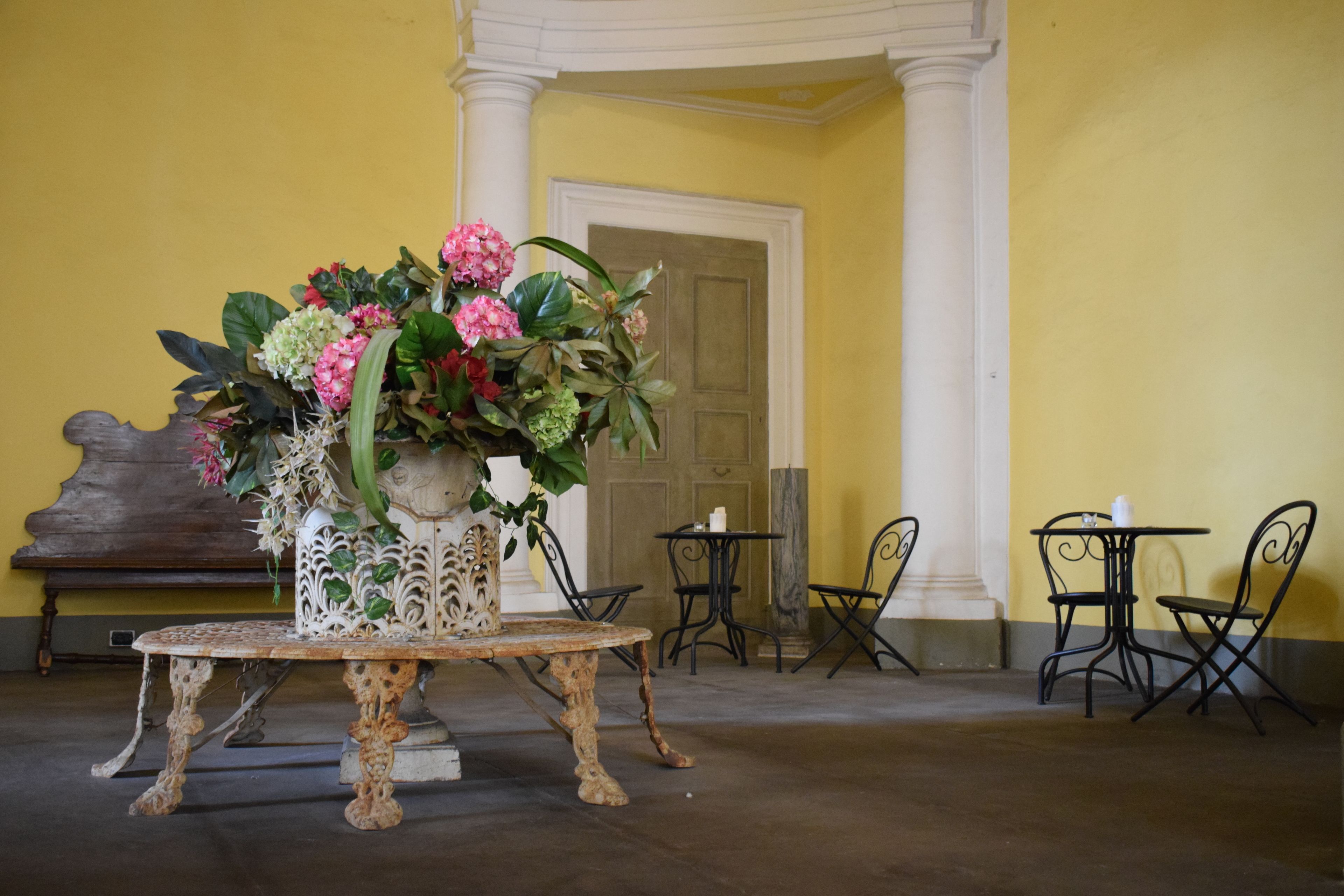 Una mesa con un arreglo floral en una habitación con paredes amarillas, un banco de madera y dos mesitas con sillas.