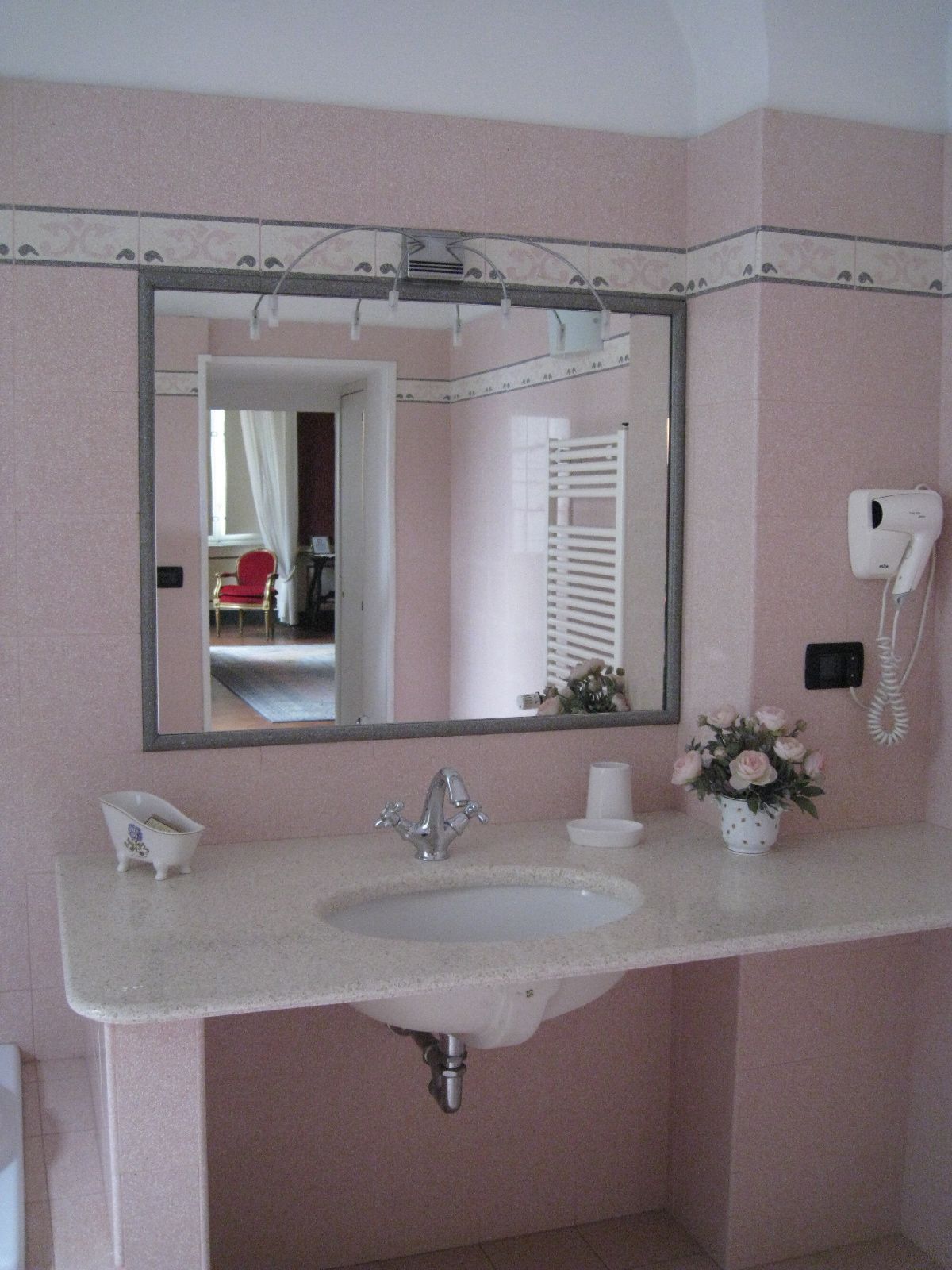 Ein Badezimmer mit einem großen Spiegel, einem Waschbecken, einem Haartrockner und einer kleinen Vase mit Blumen.