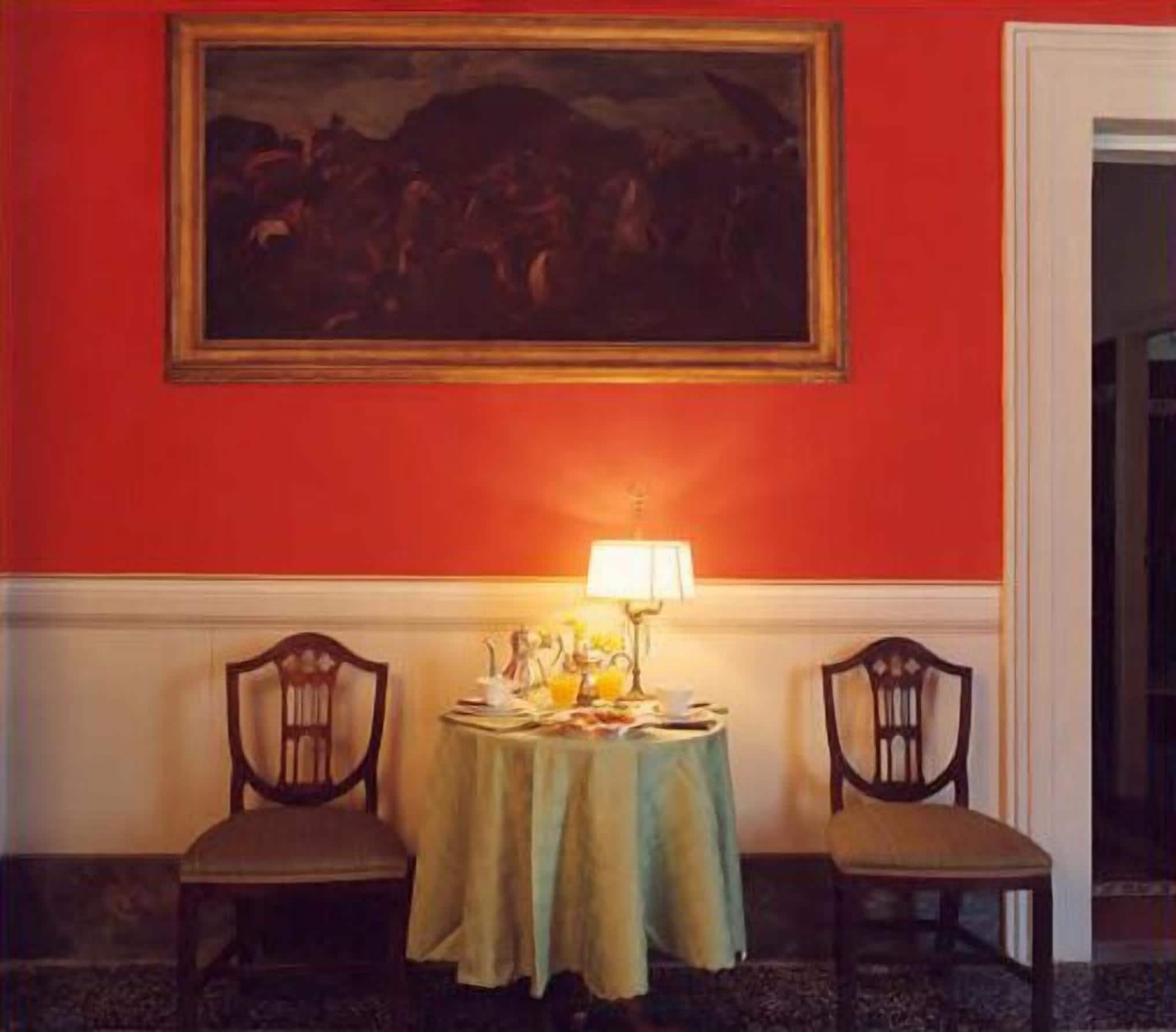 Una habitación con una pared roja, un cuadro sobre una mesa, dos sillas y una mesa con una lámpara y objetos encima.