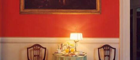 Una habitación con una pared roja, un cuadro sobre una mesa, dos sillas y una mesa con una lámpara y objetos encima.