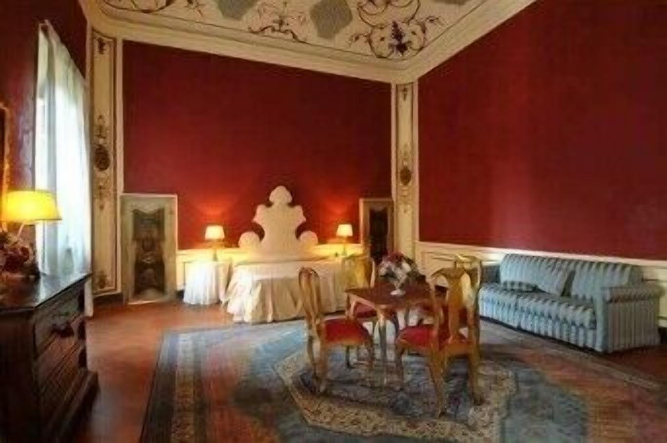 A room with a red wall, a white ornate ceiling, a dining table with chairs, a sofa, and a lamp on a side table.