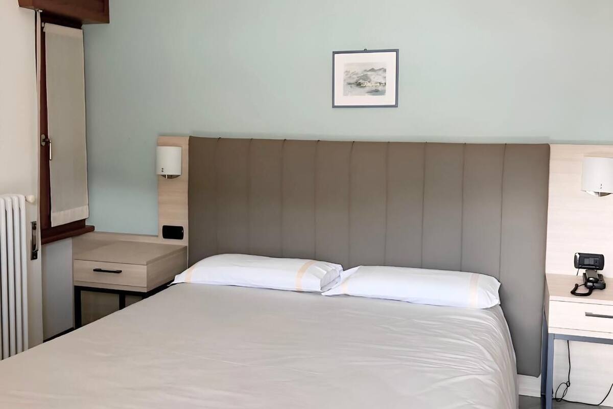 Siististi pedattu sänky hotellihuoneessa, jossa on pääty, yöpöydät lamppuineen, seinällä oleva kehystetty kuva ja patteri.