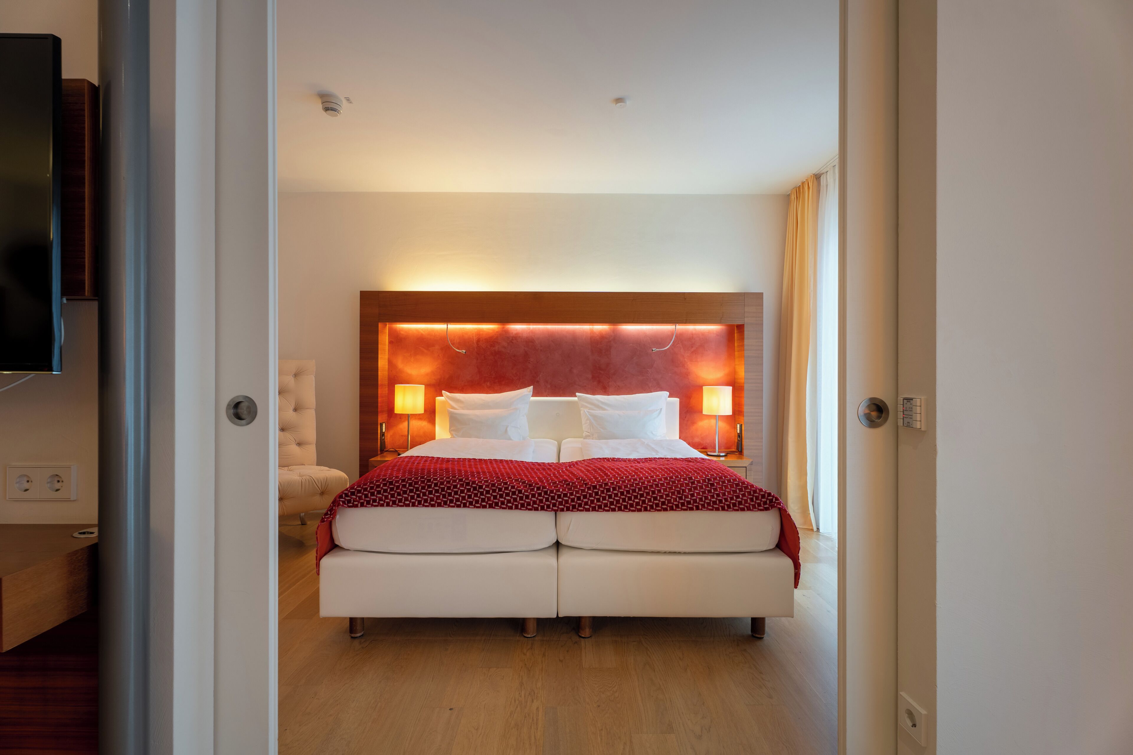 Una camera d'albergo moderna con un letto grande, lampade sui comodini, una testiera in legno e una TV a schermo piatto montata a parete.