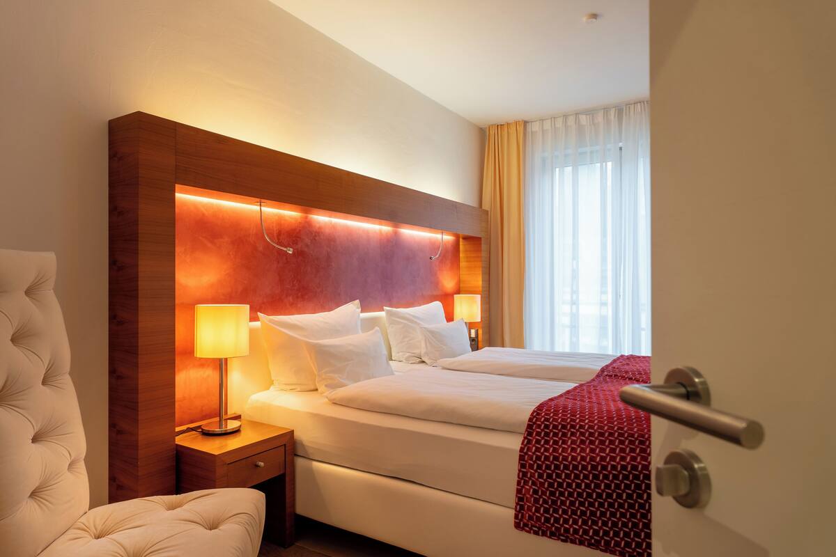 A hotel room with a large bed, a bedside table with a lamp, and a view of the outside through a window.