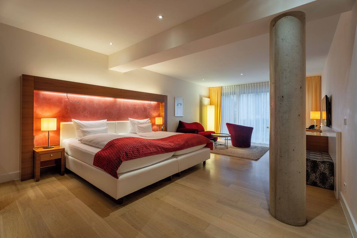 A modern hotel room with a large bed, a wooden headboard, a nightstand with a lamp, a red throw blanket, and a large pillar.