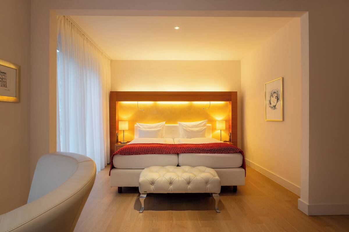A modern hotel room with a large bed, a bedside table, a chair, and a wooden floor.