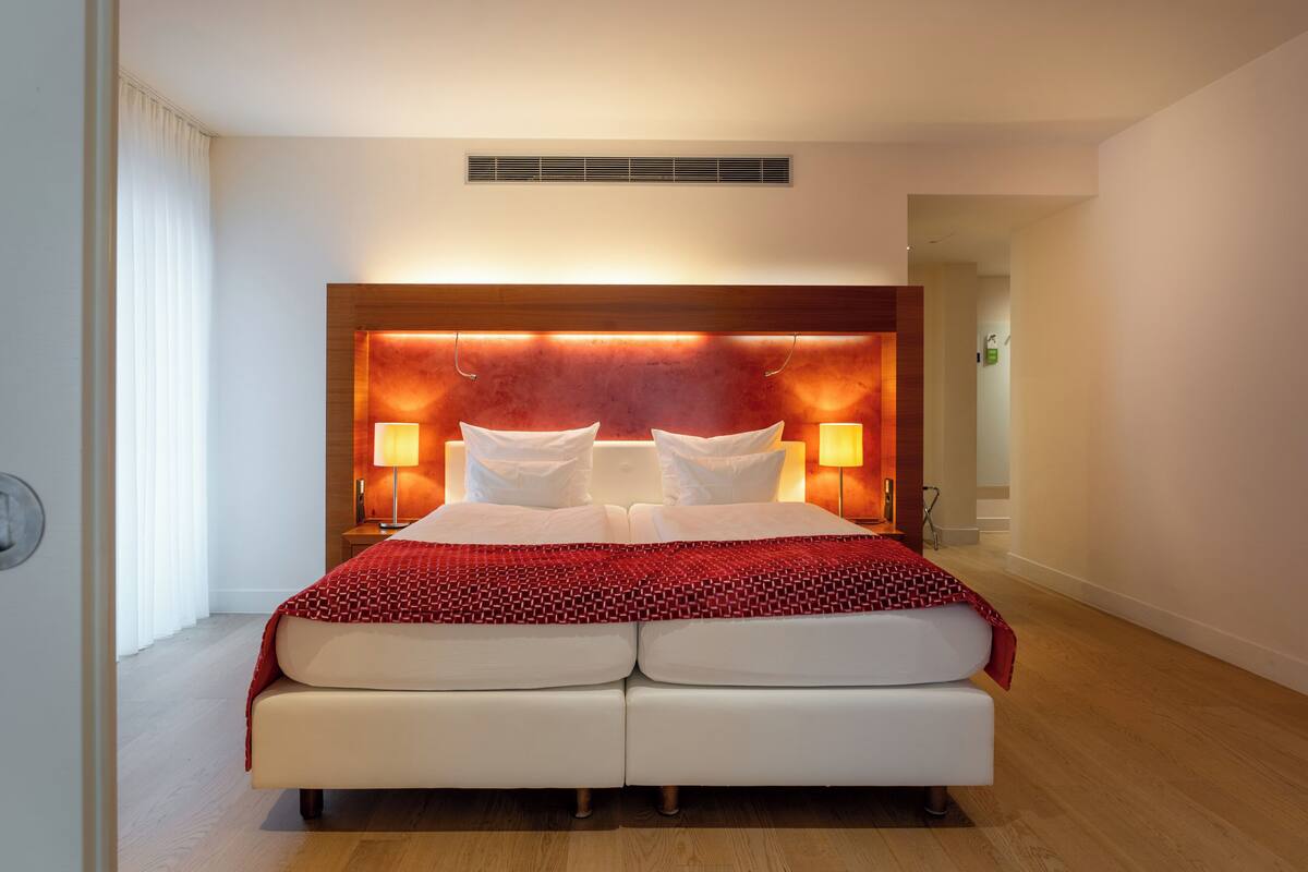 A double bed with a red patterned bedspread, two bedside lamps, and a wooden headboard with a built-in light panel.