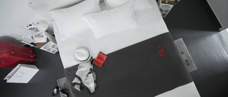 Ein ordentlich gemachtes Bett mit weißen Kissen und einer schwarz-weißen Tagesdecke, einen roten Koffer, eine weiße Lampe und ein rotes Etikett am Bett.