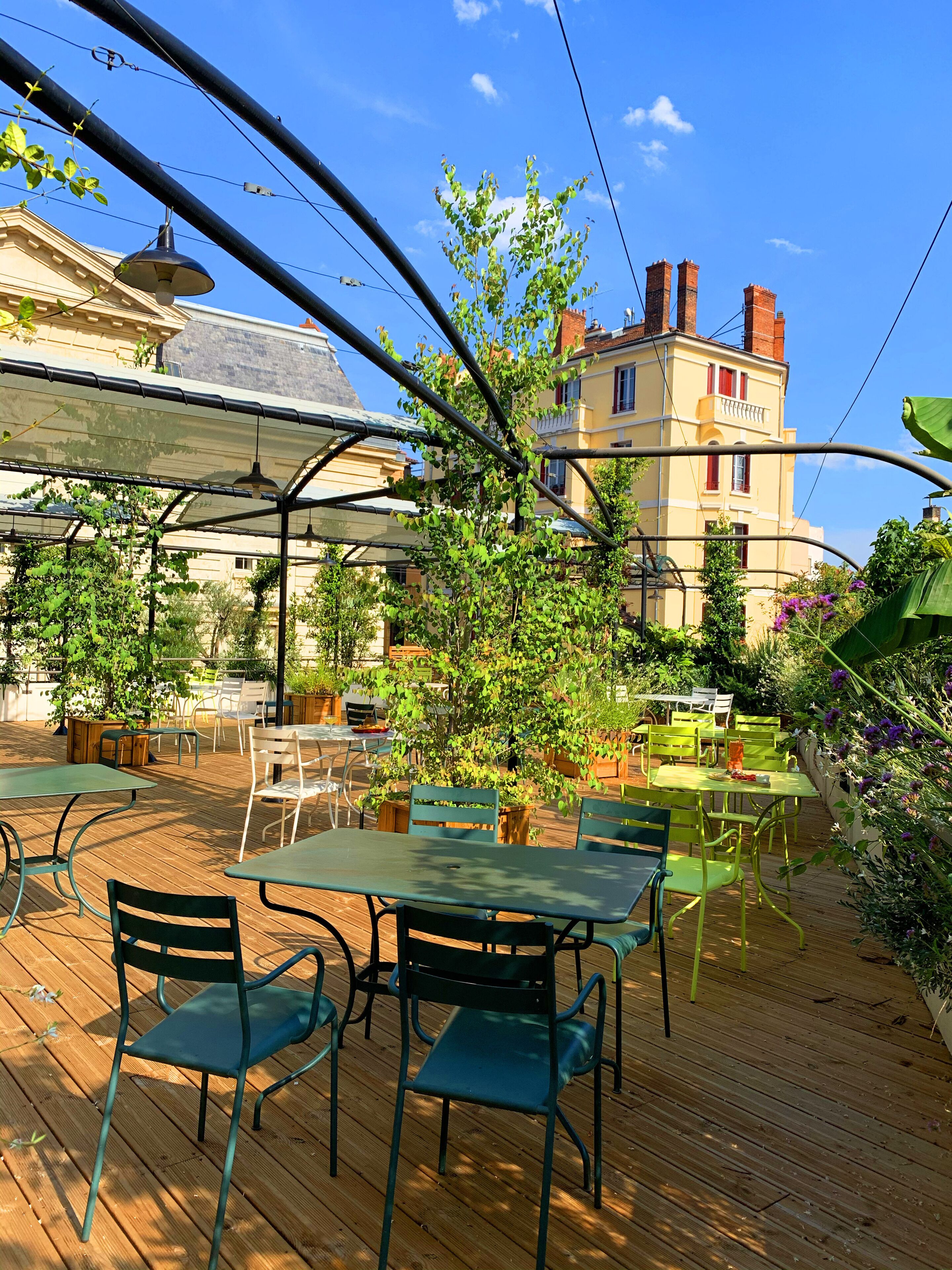 An outdoor patio with tables and chairs, surrounded by greenery and a building with red windows.