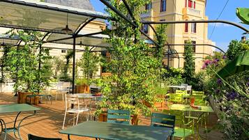 An outdoor patio with tables and chairs, surrounded by greenery and a building with red windows.