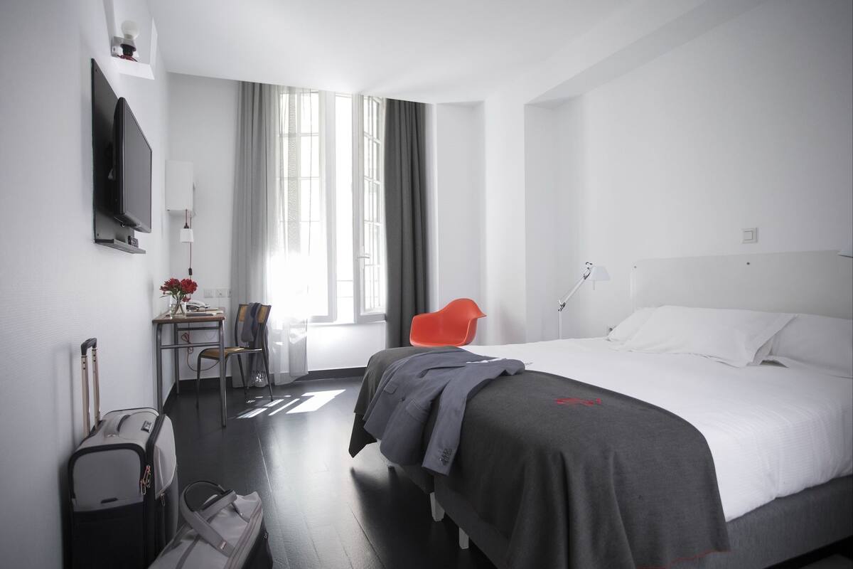 Una habitación de hotel con cama, escritorio, silla, televisión y ventana con cortinas.
