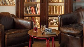 Een leren fauteuil en een klein rond tafeltje met een vintage camera en een glas. Achtergrond: een boekenplank vol boeken.