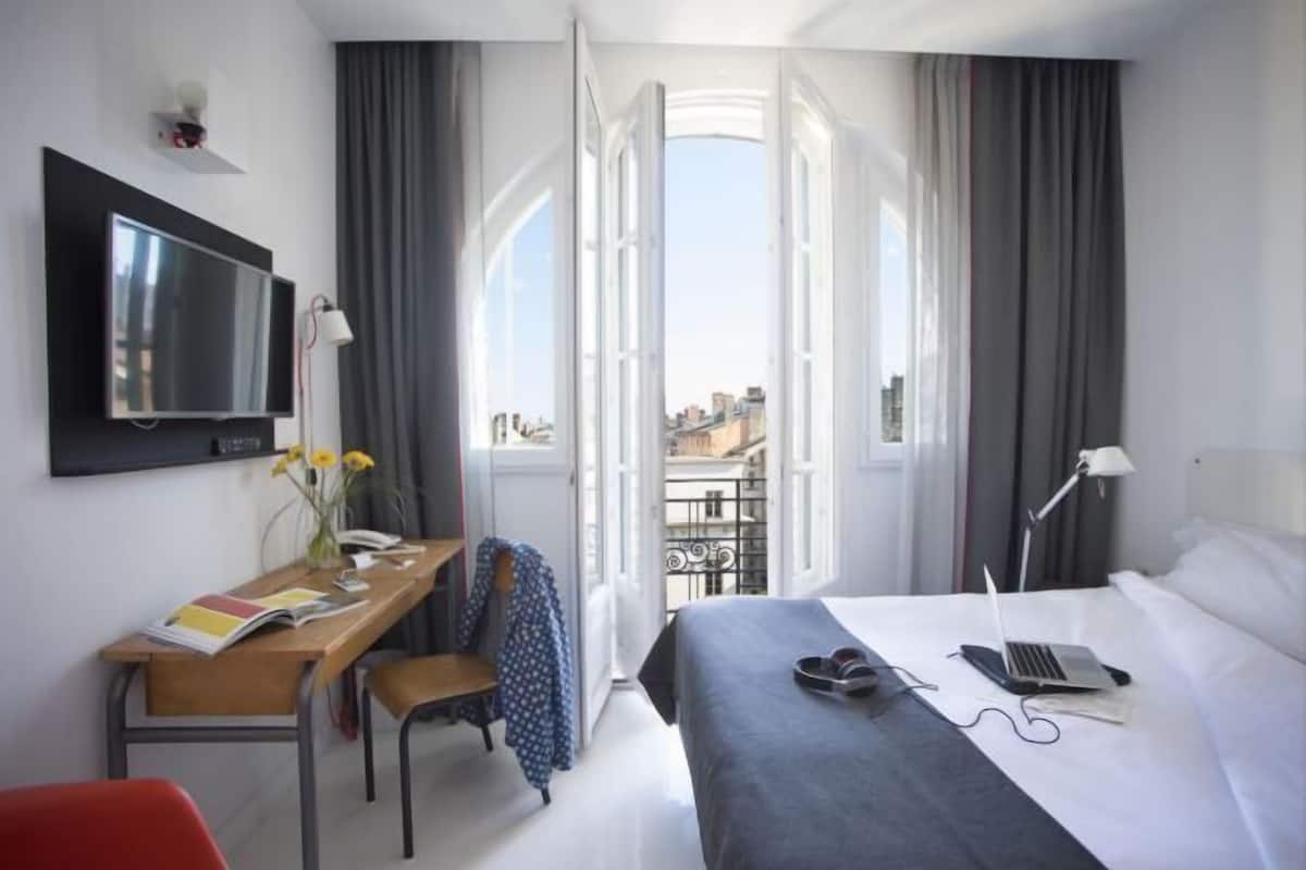 Habitación de hotel con una cama, un escritorio con silla, televisor y vistas a la ciudad a través de ventanas abiertas.