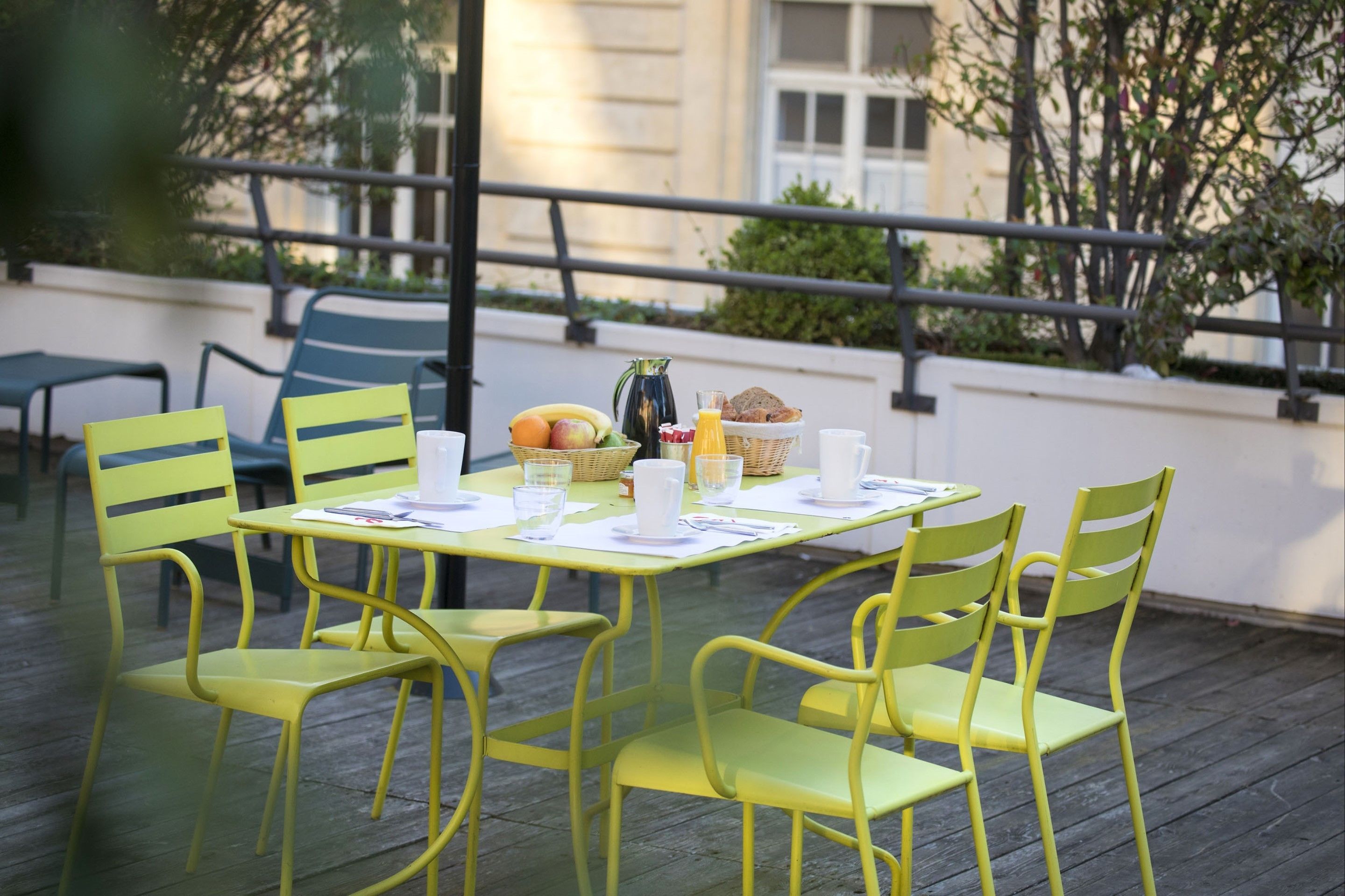 A yellow table set with chairs, a basket of fruits, a pitcher, and a cup of coffee on an outdoor patio.