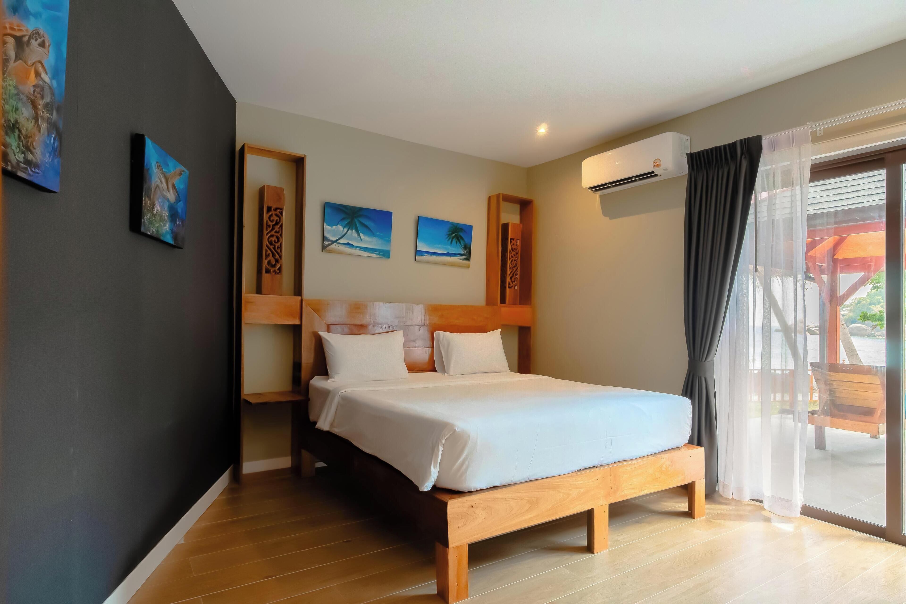A bedroom with a wooden bed, a headboard with decorative carvings, a ceiling fan, and a view of the outdoors through a large window.